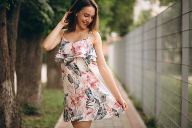 A smiling woman in a floral summer dress stands on a shaded pathway