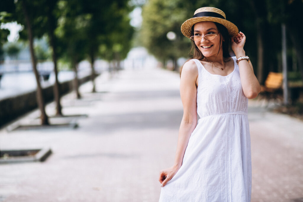 A cheerful woman in a white summer dress and straw hat stands on a tree-lined pathway